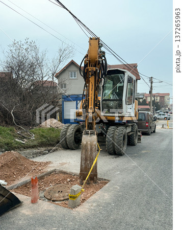 Excavator working on road repair in a suburban residential street with houses and utility lines. Urban infrastructure maintenance, municipal construction activity, civil engineering and public service Excavator working on road repair in a suburban residential street with houses and utility lines. Urban infrastructure maintenance, municipal construction activity, civil engineering and public service 137265963