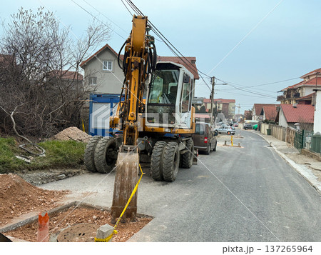 Excavator working on road repair in a suburban residential street with houses and utility lines. Urban infrastructure maintenance, municipal construction activity, civil engineering and public service Excavator working on road repair in a suburban residential street with houses and utility lines. Urban infrastructure maintenance, municipal construction activity, civil engineering and public service 137265964