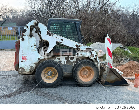 Compact skid steer loader parked on asphalt near roadwork barriers and construction materials. Urban infrastructure maintenance, heavy equipment operation and municipal engineering activity. Compact skid steer loader parked on asphalt near roadwork barriers and construction materials. Urban infrastructure maintenance, heavy equipment operation and municipal engineering activity. 137265966