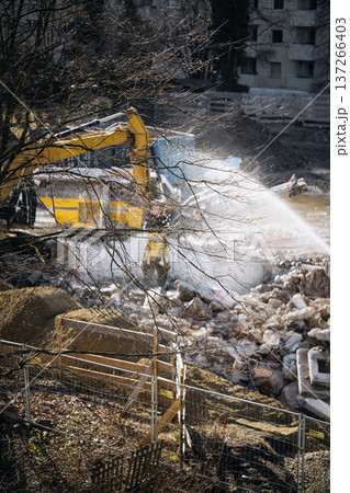Urban Building Demolition Site. Residential Construction Site Behind Apartment Windows: View Through Glass Showing Excavator, Demolition Debris, Water Spray, And Urban Renovation Activity In A 137266403