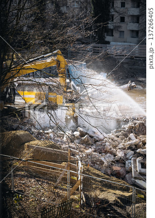 Urban Building Demolition Site. Residential Construction Site Behind Apartment Windows: View Through Glass Showing Excavator, Demolition Debris, Water Spray, And Urban Renovation Activity In A Urban Building Demolition Site. Residential Construction Site Behind Apartment Windows: View Through Glass Showing Excavator, Demolition Debris, Water Spray, And Urban Renovation Activity In A 137266405