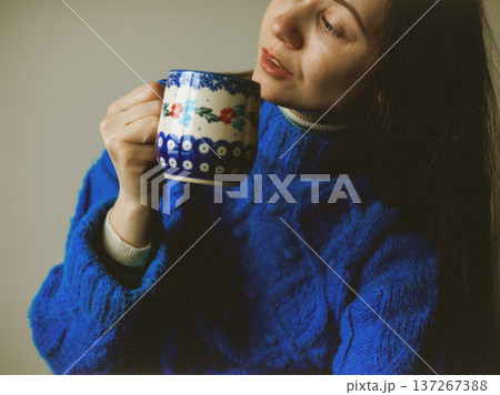 Authentic lifestyle portrait of a young woman in a vibrant cobalt blue knitted sweater holding a patterned ceramic mug. Cozy morning at home, peaceful mood, and warm indoor lighting aesthetic. 137267388