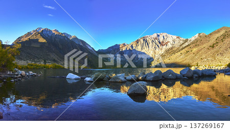 Panorama of Mountain Peaks and Clear Lake with Rocky Shore under Blue Sky Panorama of Mountain Peaks and Clear Lake with Rocky Shore under Blue Sky 137269167