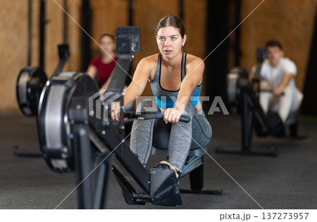 Focused girl works out with rowing machine during class at fitness club Focused girl works out with rowing machine during class at fitness club 137273957