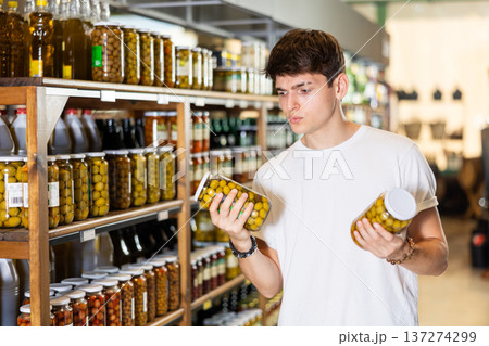 Young guy in supermarket selects examines compares two cans of olives. 137274299