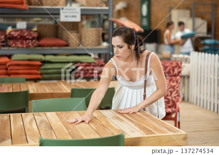 Pretty brunette girl in white dress chooses wooden table for country house in garden store Pretty brunette girl in white dress chooses wooden table for country house in garden store 137274534
