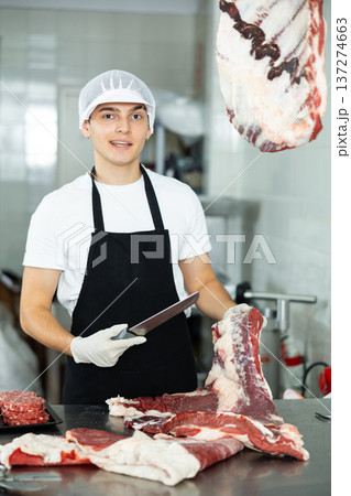 Guy employee of butcher shop cuts meat carcass into small pieces of meat for retail sale in store 137274663