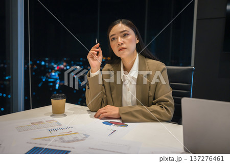 Tired businesswoman thinking and analyzing business reports late at night in office with city lights background Tired businesswoman thinking and analyzing business reports late at night in office with city lights background 137276461