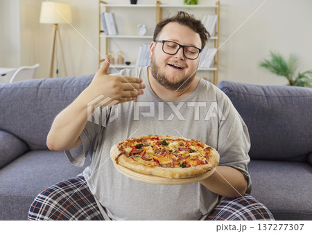 Happy man at home presenting pizza on sofa, casual pajamas, comfort moment Happy man at home presenting pizza on sofa, casual pajamas, comfort moment 137277307