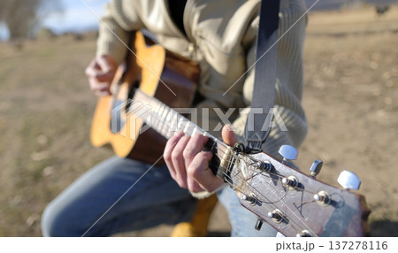 A man is playing guitar in a field. Focus on the guitar neck, strings and arm. The guitar is old and worn. Guitar lessons in the fresh air A man is playing guitar in a field. Focus on the guitar neck, strings and arm. The guitar is old and worn. Guitar lessons in the fresh air 137278116