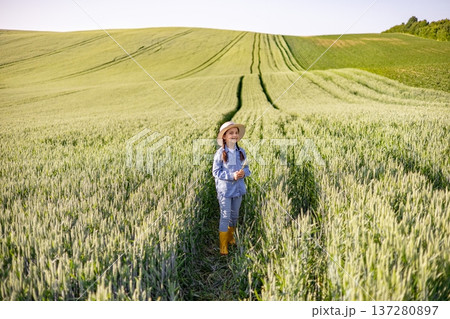 A young girl wearing a straw hat and yellow boots stands in a vast green wheat field under a clear sky A young girl wearing a straw hat and yellow boots stands in a vast green wheat field under a clear sky 137280897