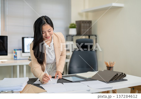 Smiling asian businesswoman or office worker holding pen over document standing leaning over table. 137284612