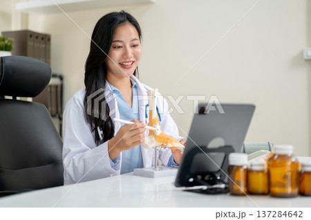 An asian woman doctor holding bone model talking on video call in tablet at table in diagnosis room. An asian woman doctor holding bone model talking on video call in tablet at table in diagnosis room. 137284642