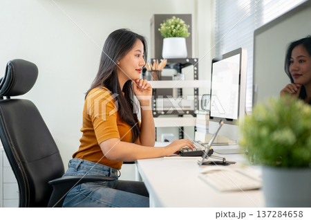 Pretty asian woman freelancer or office worker looking at computer sitting at table in company room. Pretty asian woman freelancer or office worker looking at computer sitting at table in company room. 137284658