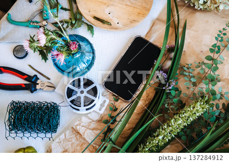 Overhead view shows smartphone on table surrounded by florist tools and botanical stems for flower arranging. Floral masterclass, botanical education, creative flower arranging course. Overhead view shows smartphone on table surrounded by florist tools and botanical stems for flower arranging. Floral masterclass, botanical education, creative flower arranging course. 137284912