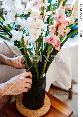 Man places gladiolus stems into glass vase on table. Seasonal flowers, locally grown blooms, gladiolus arrangement, conscious consumerism, slow flower movement. 137284914