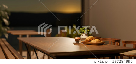 Bread and coffee with pot plant on wooden table in dim cafe dining room with sunrise outside window. Bread and coffee with pot plant on wooden table in dim cafe dining room with sunrise outside window. 137284990