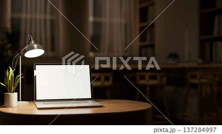 White screen laptop and lamp aside plant on round wooden table across window in dim library at night White screen laptop and lamp aside plant on round wooden table across window in dim library at night 137284997
