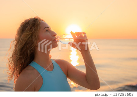 Fit woman drinking water at the beach after running outdoors in the morning. Sporty female relaxing after training. 137285608