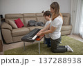 Mother guides her sons hands on a keyboard, teaching music and building warm family bonding in a relaxed home setting as they sit on the carpet practicing together 137288938