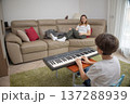 Son playing an electronic keyboard while his mother relaxes on the sofa, supporting his musical hobby and enjoying a moment of family bonding and learning in their cozy living room 137288939
