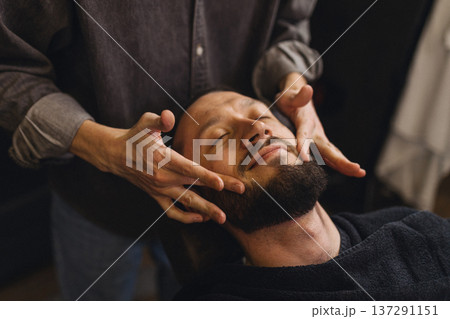 Barber massaging beard and neck of male client after shaving in barbershop. Relaxing grooming service, men self care, comfort and classic barber ritual concept. Barber massaging beard and neck of male client after shaving in barbershop. Relaxing grooming service, men self care, comfort and classic barber ritual concept. 137291151