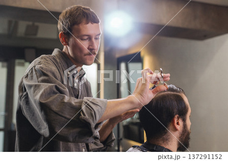 Barber cutting male client hair with scissors and comb. Close up of professional haircut process, precision technique, men grooming service in modern barbershop. Barber cutting male client hair with scissors and comb. Close up of professional haircut process, precision technique, men grooming service in modern barbershop. 137291152