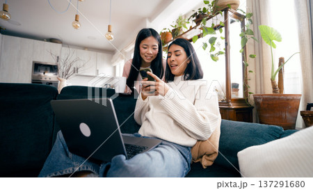 Asian women scrolling smartphone and working on laptop at home sofa Asian women scrolling smartphone and working on laptop at home sofa 137291680