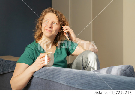 Young happy woman enjoying sunny morning at home, sipping coffee on the sofa, smiling and chatting on smartphone 137292217