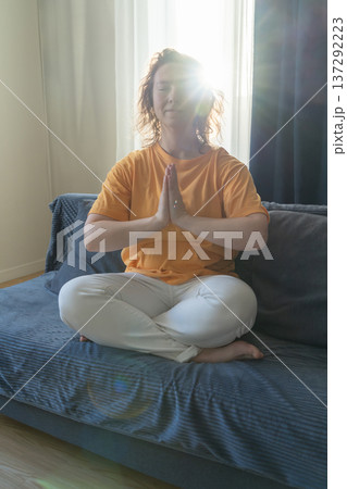 Woman sits in lotus position with her hands folded in prayer on sofa under bright ray of sunlight, practicing meditation and finding inner peace at home Woman sits in lotus position with her hands folded in prayer on sofa under bright ray of sunlight, practicing meditation and finding inner peace at home 137292223
