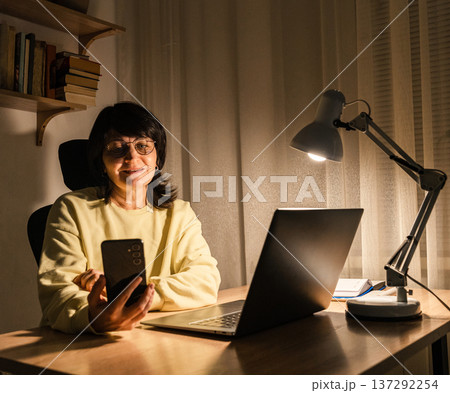 Mature woman in glasses working from home night, smiling while using smartphone laptop at desk with lamp light, working remotely Mature woman in glasses working from home night, smiling while using smartphone laptop at desk with lamp light, working remotely 137292254