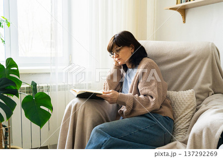 Woman in glasses relaxing in an armchair by the window, engrossed in a book, enjoying leisure time at home Woman in glasses relaxing in an armchair by the window, engrossed in a book, enjoying leisure time at home 137292269