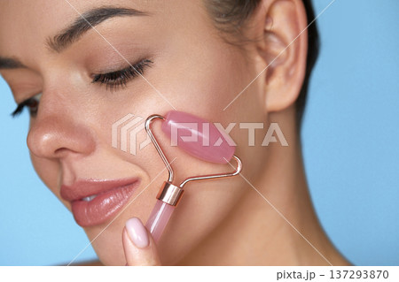 Woman using jade facial roller for face massage. Girl using natural massager tool for skin care and spa beauty treatment closeup portrait. 137293870