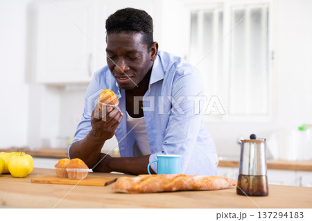 Positive African American eating muffin leaning on countertop in kitchen Positive African American eating muffin leaning on countertop in kitchen 137294183