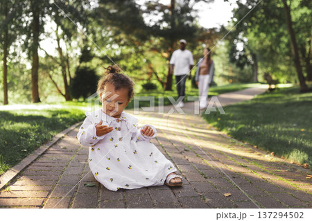 Curious little daughter sitting on park path and exploring nature while parents walk behind. Family lifestyle outdoors, childhood independence, care and safety. Small child at summer day in green park Curious little daughter sitting on park path and exploring nature while parents walk behind. Family lifestyle outdoors, childhood independence, care and safety. Small child at summer day in green park 137294502