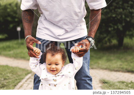 Happy little daughter smiling while father holds hands and helps walk in park. Joyful family outdoors, parenting, trust and support, love and development in nature. Dad and girl child Happy little daughter smiling while father holds hands and helps walk in park. Joyful family outdoors, parenting, trust and support, love and development in nature. Dad and girl child 137294505
