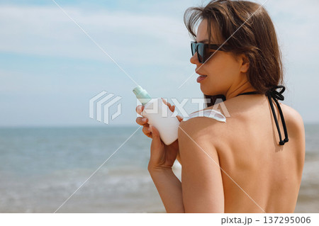 Beautiful female model holding sunscreen bottle and applying suncream on shoulder at seaside, summer skincare and UV protection concept with healthy glowing skin 137295006
