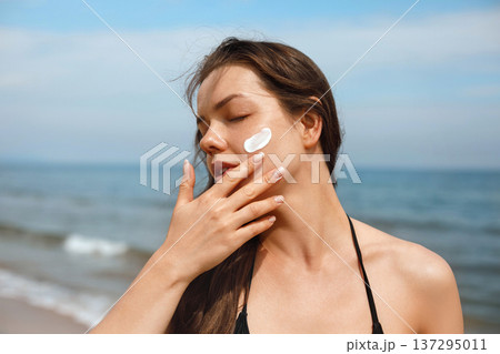 Attractive female applying sunscreen lotion on face with closed eyes at seaside, beauty skincare and sun protection concept with relaxed summer mood Attractive female applying sunscreen lotion on face with closed eyes at seaside, beauty skincare and sun protection concept with relaxed summer mood 137295011