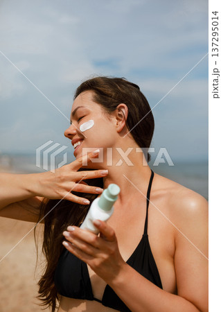 Attractive young woman holding sunscreen bottle and touching face at beach, skincare routine and sun protection concept with fresh healthy skin Attractive young woman holding sunscreen bottle and touching face at beach, skincare routine and sun protection concept with fresh healthy skin 137295014