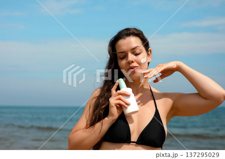 Close portrait of woman applying sunscreen suncream lotion on cheek at beach, female using sunblock for skin care and UV protection 137295029