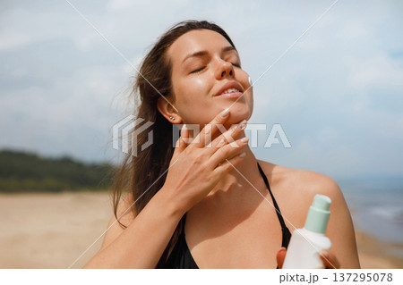 Young woman gently applying sunscreen lotion on cheek at beach under sunlight, facial skincare routine and sun protection concept with natural beauty 137295078