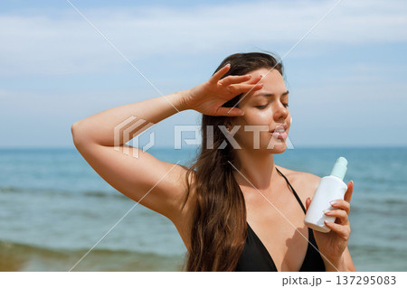 Young woman holding sunscreen bottle and shielding eyes from sun on beach using suncream lotion for UV protection and skincare routine Young woman holding sunscreen bottle and shielding eyes from sun on beach using suncream lotion for UV protection and skincare routine 137295083