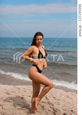 Slim young woman standing on beach in bikini posing with confidence enjoying sun and applying sunscreen suncream lotion for skin protection and summer body care routine 137295098
