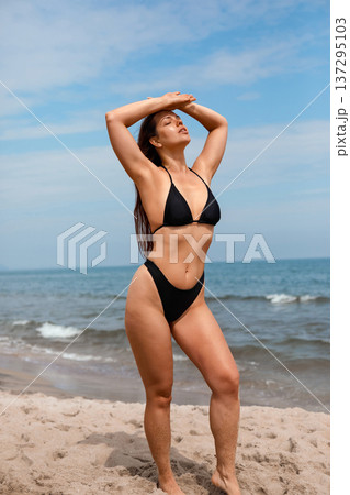 Young woman standing on beach with hands above head enjoying sun and applying sunscreen suncream lotion for skin protection and summer skincare routine 137295103