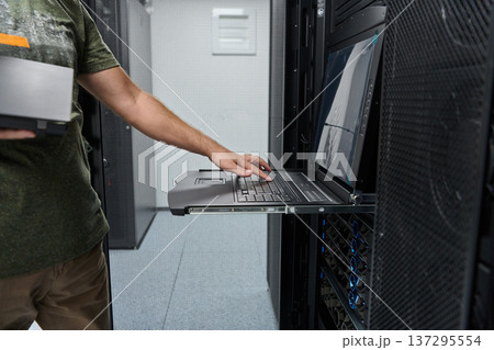 It technician working on a laptop console in a data center server rack performing maintenance and monitoring It technician working on a laptop console in a data center server rack performing maintenance and monitoring 137295554