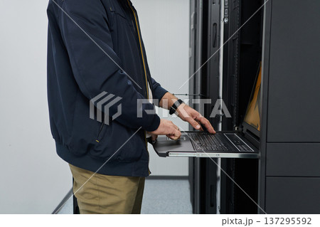 IT technician working on laptop in server rack inside data center performing network maintenance and hardware monitoring 137295592