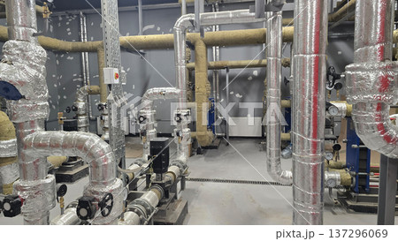 Industrial piping system featuring various insulated pipes, valves, and control panels arranged in mechanical room, showcasing clean and organized layout with metallic surfaces and functional design 137296069