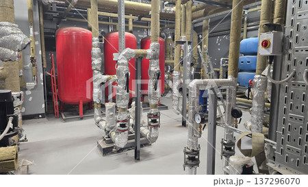 Industrial piping system featuring insulated pipes, large red and blue tanks, and various valves and gauges within a mechanical room, showcasing a complex network of equipment and infrastructure Industrial piping system featuring insulated pipes, large red and blue tanks, and various valves and gauges within a mechanical room, showcasing a complex network of equipment and infrastructure 137296070