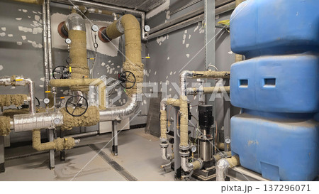 Industrial plumbing setup with insulated pipes and valves connected to a large blue water tank in a utility room, showcasing gray walls with visible wear and maintenance signs Industrial plumbing setup with insulated pipes and valves connected to a large blue water tank in a utility room, showcasing gray walls with visible wear and maintenance signs 137296071