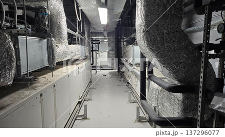 Industrial corridor showcasing extensive metallic ductwork and ventilation systems along ceiling, with white walls and flooring, illuminated by overhead lights in commercial building environment Industrial corridor showcasing extensive metallic ductwork and ventilation systems along ceiling, with white walls and flooring, illuminated by overhead lights in commercial building environment 137296077